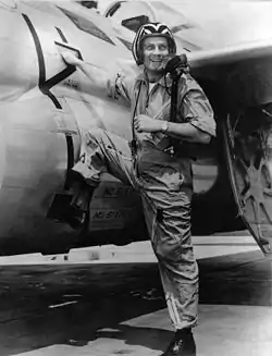 A monochrome photograph of an energetic man in his 30s wearing a jet pilot uniform and helmet, stepping with one booted foot up onto a military jet, his right hand on the jet, turning back to smile at the viewer