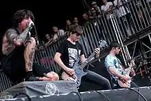 Three men performing on stage. One is kneeling on one knee and singing into a microphone while the other are next to him playing the guitar.