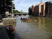 A yellow water taxi on the water between stone quaysides. The far bank has large buildings and in the distance is a three arch bridge.