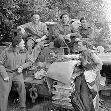 Three soldiers sit on a tank while two more stand next to it.