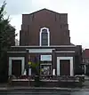 A plain building in very dark brick. In the foreground is a flat-roofed entrance area with two wooden doors and a small window (partly obscured).  Behind this, a squat tower, with a central window, rises in several stages.