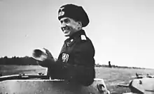 A smiling Munzel wearing a black military uniform stands inside his tank turret.