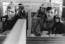 A black and white picture of two students wearing headphones. One student's booth is numbered "17", and the other's is numbered "18".