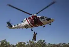 A helicopter painted red and white flying with two people wearing military uniforms hanging from a rope attached to the helicopter