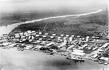 Black and white photo of an industrial installation located next to a body of water. It includes a large number of white storage tanks and buildings, as well as a wharf. A large ship is docked at the wharf.