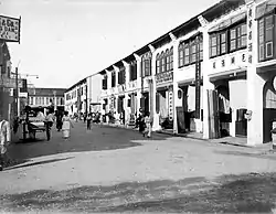 High angle view of a street lined with two and three-story structures. The street is filled with crowds, including horse-drawn and hand-pulled carriages.