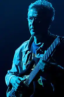 A white man with gray hair, wearing a striped shirt with white and pink colors, holding an acoustic guitar with two microphones in front of him.