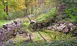 Two intersecting stone walls below a larger stone retaining wall in a grassy, shrubby area. There is a small set of table and chairs next to a large vase in a cleared brick area at the corner