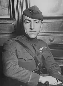 Head and torso of a man turned in his chair, his right arm lying stiffly on the arm rest in front of him. He is wearing a military uniform with a strap diagonally across the chest, a winged emblem on his left breast, and a garrison cap.