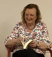 A smiling white mature woman with wavy light brown hair, with glasses hanging on a cord round her neck, sits holding an open book