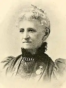 An older white woman, with white hair, wearing a high lace collar and large sleeves; she has a pin or medallion on her lapel.