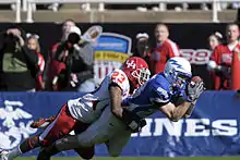 An American football in a white jersey tackles one in a blue jersey catching a ball.