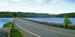 Ground-level view of a causeway crossing a reservoir. On either side of the causeway are short stone walls. The lake's banks are surrounded by thick woods.