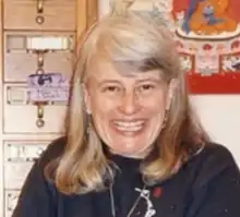 tight headshot of a grinning white older woman with shoulder length hair standing on front of a card catalog