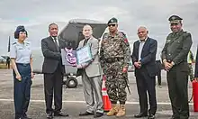 Colour photo of seven people wearing a mix of military and business attire posing in front of an aircraft