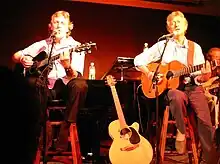 Chad and Jeremy seated on stools, singing and playing guitars