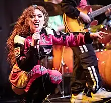 Chappell Roan crouching onstage in a pink and black jumpsuit. She is holding a microphone and pointing.