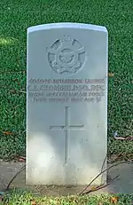 A white grave stone marker. The stone contains an insignia at the top with a crown, flying eagle and banners, followed by text of the person and a Christian cross.