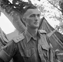 an informal portrait of a bare-headed man dressed in uniform with tents behind him