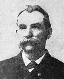 Head of a white man with a large drooping mustache and receding hairline, wearing a dark suit coat over a shirt and tie.