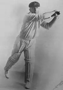 Studio portrait against a white background of a stocky cricketer in white shirt and trousers with rolled up sleeves, cricket pads, baggy green cap with Australia coat of arms, wearing gloves and holding a bat and swinging it horizontally to his left. He is a right-hander and his leading left leg is straight but his right leg is bent with only the toes on the ground.