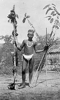 Black and white photograph of a man in a loincloth and feathers with a pole in one hand and four long sticks in the other.