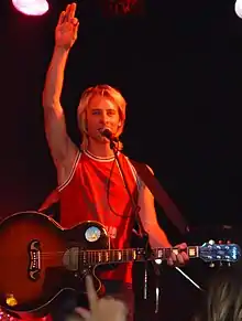 A white man in his mid-thirties stands on a stage. He is reasonably tall, and has light brown hair with blonde highlights. He is holding a microphone in front of his mouth, and has a guitar in his hand. He is wearing jeans and a red top.