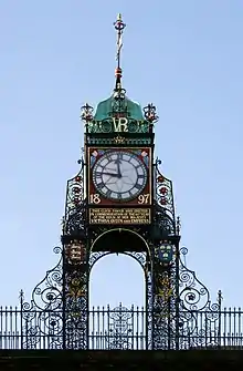 A highly ornate structure in wrought iron, with a railing at the base.  It carries a clock with Roman numerals; this has a red frame bearing the date "1897" in gold, and under it is a plate with an inscription in gold lettering.  Above the clock are the initials "VR" in gold, and at the summit is a green cupola with a weather vane.