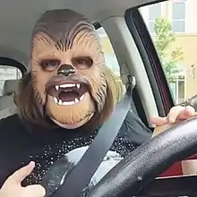 A woman wearing a Chewbecca mask and a Death Star t-shirt looks into the camera from the driver's seat of her car