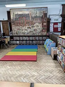 Area of a library with wooden shelves, illustration of children playing and coloured mats on the floor