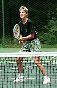 A blonde-haired female tennis player with multi-colored shorts and a black shirt, with the tennis racket out in front of her