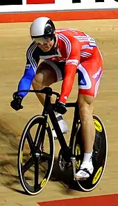 Chris Hoy wearing a bicycle helmet, visor, cycling shorts and top cycling on a racing bike in a velodrome.