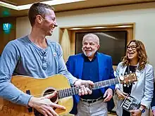 A casually-dressed man holding a guitar smiles along with another man and a woman, who are dressed formally