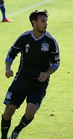 A man wearing a black jersey and black shorts, running on a soccer field.