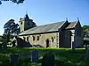 A small church seen from the southeast. The tower has a small pyramidal spire, a porch protrudes from the south wall, and the chancel to the right is short and slightly lower than the nave. Gravestones in shadow are in the foreground