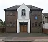 A two-storey building with a dark brick façade dominated by a central gabled stuccoed section. This has recess for the wooden entrance door and an arched window.  Both storeys have six small windows with blue frames.