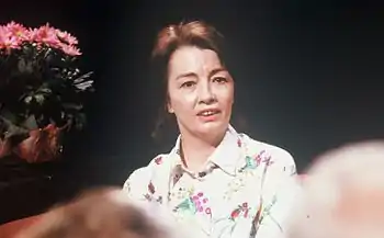 A photograph of a woman with shoulder-length brown hair facing the viewer and looking slightly to the right while wearing a white shirt with a floral print