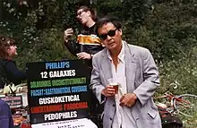 Chu stands next to a sign that reads "Phillips / 12 Galaxies / Solarunikel unconstitutionality / Pacsat: Kastronoticul coverage / Guskoketical / Libertarians parochial / Pedophiles"