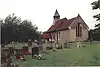 A small stone church with red tiled roofs seen through a churchyard from the southeast. On its far gable is a bellcote with a pyramidal roof