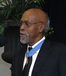 Clarence Sasser, a gray-haired black man standing at a microphone, wearing a medal on a blue ribbon around his neck.