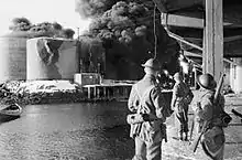 men n quayside looking across water at burning tanks