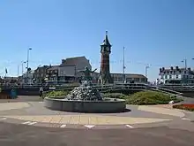 A fountain with a sculpture of a person; a clock tower, people and buildings are in the background