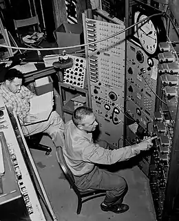 black-and-white side view of Clyde Cowan, seated with his right arm outstretched toward one of several equipment panels before him