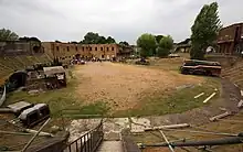 Wide view of the fort's interior, showing a semi-circular sandy area in which items of military equipment are standing. A row of brick buildings is visible at the rear.