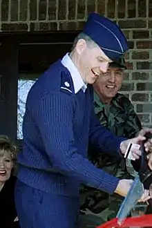 A profile photo of white man in a blue US Air Force uniform.  He is facing the right, smiling, and wielding large scissors for a ribbon-cutting ceremony.