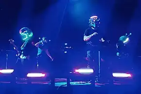 Four men wearing custom helmets perform on stage, from left to right, they are on the bass, dancing, electronic drum pad and guitar