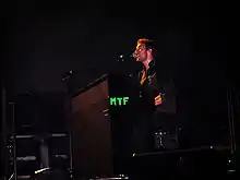 A short-haired man wearing a black outfit performs with a microphone and a piano