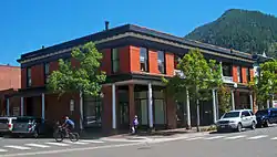 A two-story brick building on a street corner seen from the opposite corner. It is lit by the sun from the right. At the street the sidewalk is sheltered by a roof supported by round white columns; the building's exterior there is faced in rough stone. Medium-sized trees are planted along the street. At the flat roofline is a wide black and white cornice; in the background at right is a wooded mountaintop.