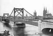 Black-and-white photograph of the Deutz Suspension Bridge in 1925. The photo was taken from the shore. One end and both towers of the bridge are visible, with the river seen flowing underneath. The Cologne cathedral is visible on the far shore.