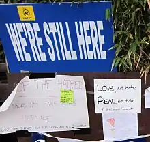 Signs reading "We're still here", "Love, not hate / Real, not fake" and other messages